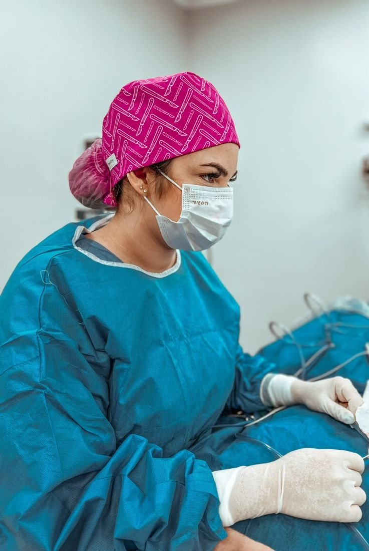 Dra. Kamila paramentada com touca rosa e avental cirúrgico durante um procedimento.