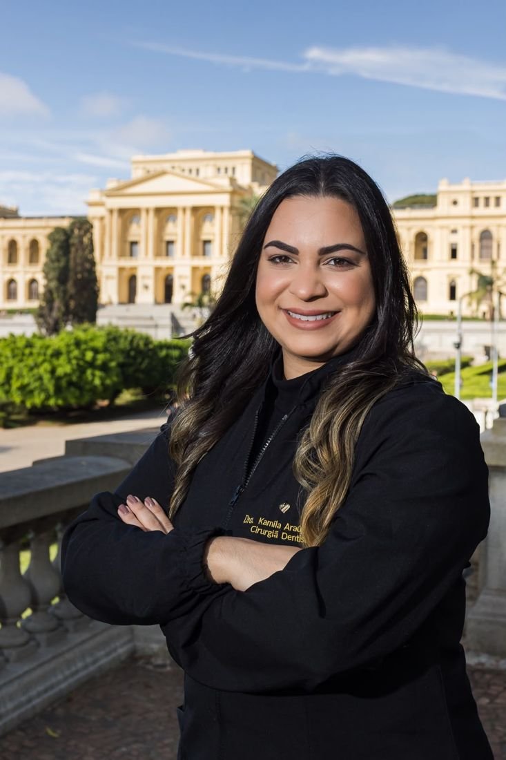 Dra. Kamila Carazzatto sorrindo, de jaleco preto, em frente ao Museu do Ipiranga.